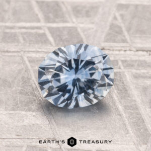 A 1.27-carat heated Montana sapphire with a blue hue is displayed on a textured silver surface. "EARTH'S TREASURY" is shown at the bottom.