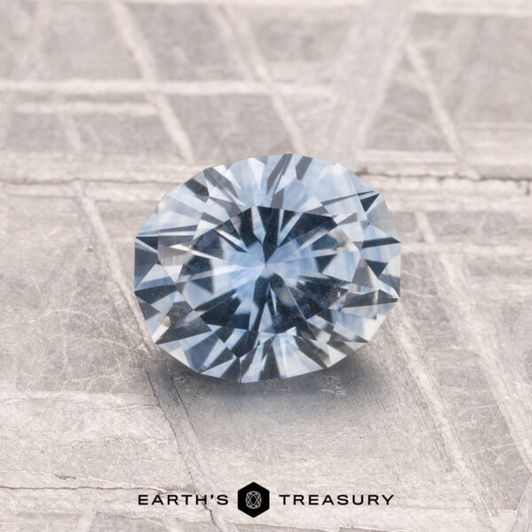 1.27-Carat Montana Sapphire (Heated) A 1.27-carat heated Montana sapphire with a blue hue is displayed on a textured silver surface. "EARTH'S TREASURY" is shown at the bottom.
