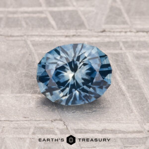 A 1.25-carat heated Montana sapphire, faceted in an oval shape and blue in color, is displayed on a textured gray surface with "EARTH'S TREASURY" at the bottom.