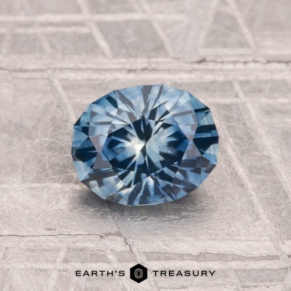 1.25-Carat Montana Sapphire (Heated) A 1.25-carat heated Montana sapphire, faceted in an oval shape and blue in color, is displayed on a textured gray surface with "EARTH'S TREASURY" at the bottom.