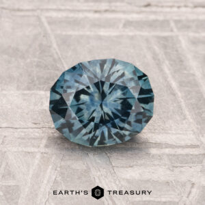 A round, faceted 1.22-carat heated Montana sapphire with a blue hue rests on a textured light gray surface. The text "EARTH'S TREASURY" appears at the bottom of the image.