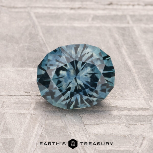 1.22-Carat Montana Sapphire (Heated) A round, faceted 1.22-carat heated Montana sapphire with a blue hue rests on a textured light gray surface. The text "EARTH'S TREASURY" appears at the bottom of the image.