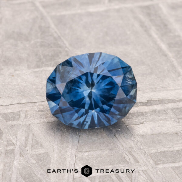 1.19-Carat Montana Sapphire (Heated) A faceted round 1.19-carat heated Montana sapphire is displayed on a textured light gray surface with "EARTH'S TREASURY" and a hexagon logo at the bottom.