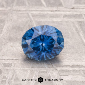 A 1.12-carat heated Montana sapphire in round brilliant cut is showcased on a textured gray surface with "EARTH'S TREASURY" at the bottom.
