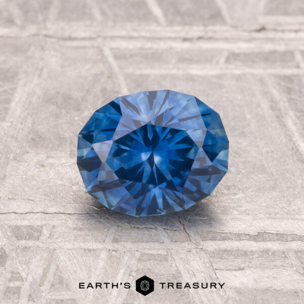 1.12-Carat Montana Sapphire (Heated) A 1.12-carat heated Montana sapphire in round brilliant cut is showcased on a textured gray surface with "EARTH'S TREASURY" at the bottom.
