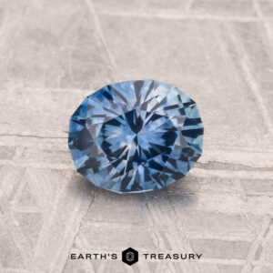 A 1.07-carat heated Montana Sapphire with a round brilliant cut is displayed on a textured gray surface, featuring the text "EARTH'S TREASURY" at the bottom.