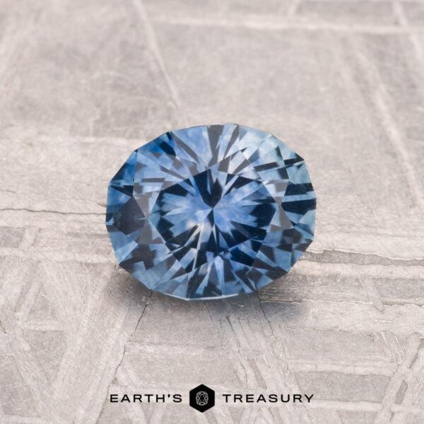 1.07-Carat Montana Sapphire (Heated) A 1.07-carat heated Montana Sapphire with a round brilliant cut is displayed on a textured gray surface, featuring the text "EARTH'S TREASURY" at the bottom.