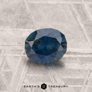 A 0.95-carat heated Montana sapphire with a round, faceted cut rests on a textured, light-colored surface. The "EARTH'S TREASURY" logo and text appear at the bottom of the image.