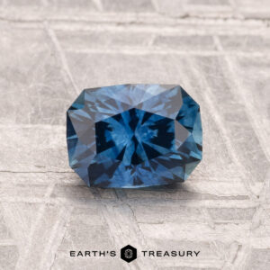A 1.25-carat heated Montana sapphire, rectangular and deep blue with faceted cuts, is displayed on a textured gray surface featuring the "EARTH'S TREASURY" text at the bottom.