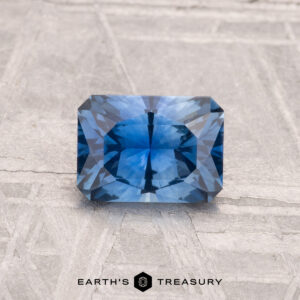 A 1.10-carat Montana sapphire (heated) with faceted edges is displayed on a textured light gray surface, featuring the text "EARTH'S TREASURY" at the bottom.