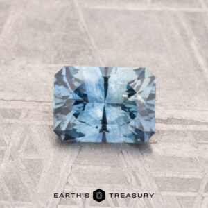 A rectangular, faceted 1.10-carat heated Montana sapphire gemstone rests on a textured gray surface, with the "EARTH'S TREASURY" logo and text at the bottom.