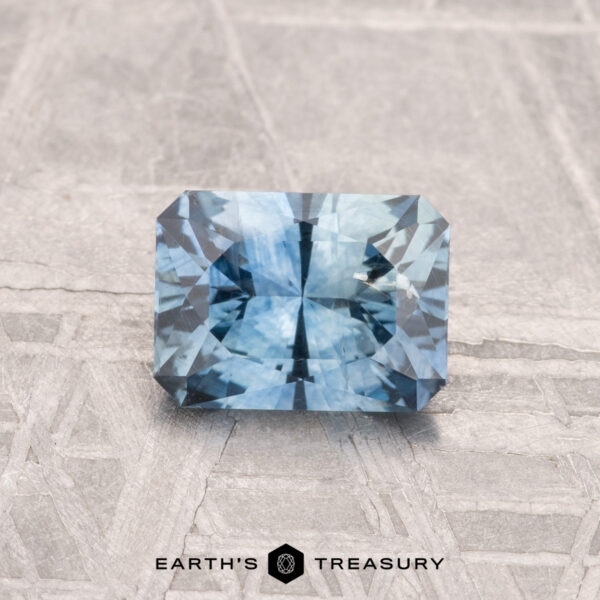 1.10-Carat Montana Sapphire (Heated) A rectangular, faceted 1.10-carat heated Montana sapphire gemstone rests on a textured gray surface, with the "EARTH'S TREASURY" logo and text at the bottom.
