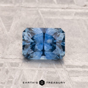 A 1.07-carat heated Montana sapphire with intricate facets rests on a textured gray surface, accompanied by the words "EARTH'S TREASURY" below it.