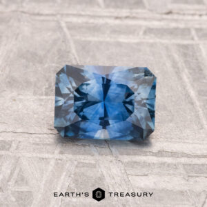A 1.02-carat heated Montana sapphire, rectangular and faceted with a blue hue, rests on a textured surface. "EARTH'S TREASURY" appears at the bottom of the image.