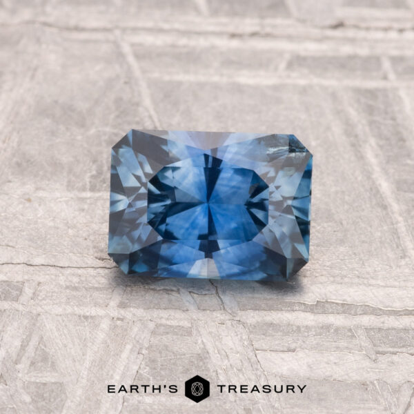 1.02-Carat Montana Sapphire (Heated) A 1.02-carat heated Montana sapphire, rectangular and faceted with a blue hue, rests on a textured surface. "EARTH'S TREASURY" appears at the bottom of the image.