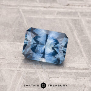 A rectangular, blue faceted 0.93-carat Montana Sapphire (Heated) is displayed on a textured gray surface with the "Earth's Treasury" logo below.