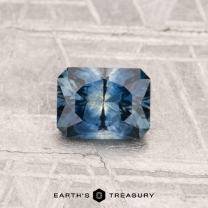 A 0.89-carat heated Montana sapphire with a rectangular cut and starburst pattern is shown on a textured gray surface. "EARTH'S TREASURY" is displayed at the bottom of the image.