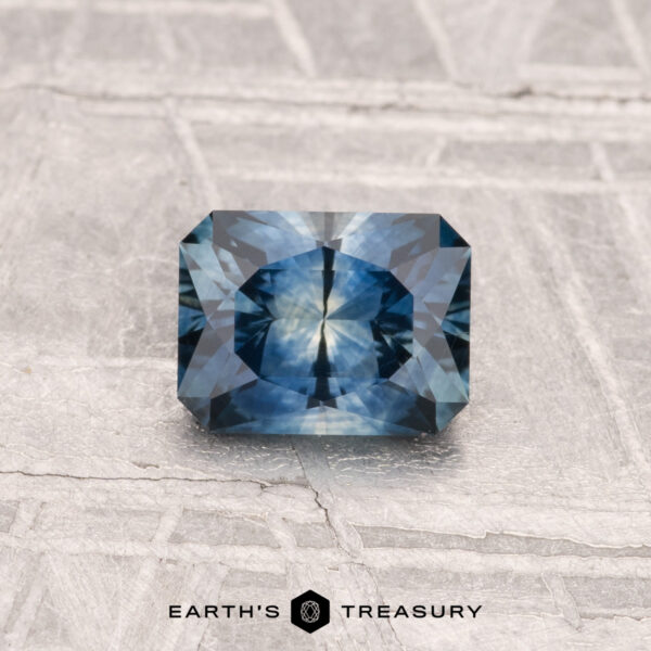 0.89-Carat Montana Sapphire (Heated) A 0.89-carat heated Montana sapphire with a rectangular cut and starburst pattern is shown on a textured gray surface. "EARTH'S TREASURY" is displayed at the bottom of the image.