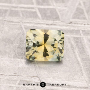 A square-cut, yellow-green 0.82-carat Montana Sapphire (Heated) is displayed on a textured gray surface with "Earth's Treasury" written below.