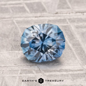 A 1.44-carat heated Montana sapphire with a radiant cut is displayed on a textured light gray surface. The "EARTH'S TREASURY" logo appears at the bottom.