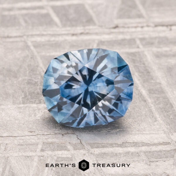 A 1.44-carat heated Montana sapphire with a radiant cut is displayed on a textured light gray surface. The "EARTH'S TREASURY" logo appears at the bottom.