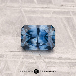 A rectangular, multi-faceted blue 0.72-carat heated Montana sapphire rests on a textured, light surface. "Earth's Treasury" appears at the bottom of the image.