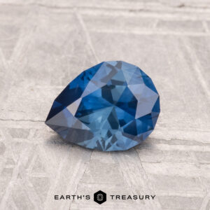 A 1.10-carat faceted, pear-shaped blue Montana sapphire (heated) is displayed on a light textured surface with the "Earth's Treasury" logo at the bottom.