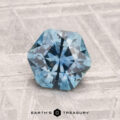 A 1.39-carat heated Montana sapphire with a faceted hexagonal cut rests on a textured gray surface, featuring the words "EARTH'S TREASURY" below.