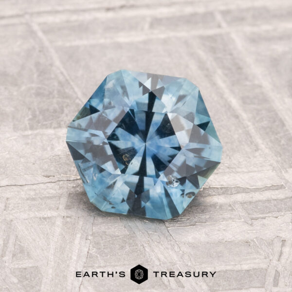 A 1.39-carat heated Montana sapphire with a faceted hexagonal cut rests on a textured gray surface, featuring the words "EARTH'S TREASURY" below.