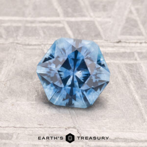 A 1.24-carat heated Montana sapphire, faceted in an octagonal shape, rests on a textured gray surface. The "Earth's Treasury" logo and text are visible at the bottom.