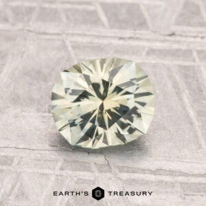 A faceted, pale green 1.34-carat Montana Sapphire (Heated) rests on a textured light gray surface featuring the "EARTH'S TREASURY" text and logo below.
