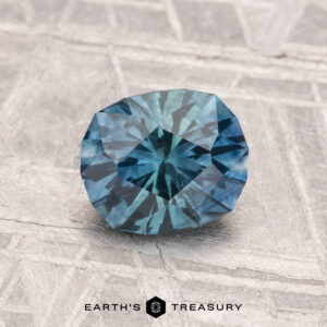 A 1.32-carat heated Montana sapphire with blue-green facets rests on a textured gray surface with the text "EARTH'S TREASURY" at the bottom.