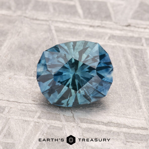 1.32-Carat Montana Sapphire (Heated) A 1.32-carat heated Montana sapphire with blue-green facets rests on a textured gray surface with the text "EARTH'S TREASURY" at the bottom.