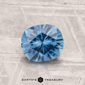 A 1.25-carat heated Montana sapphire is displayed on a textured gray surface, with the text "EARTH'S TREASURY" beneath it.