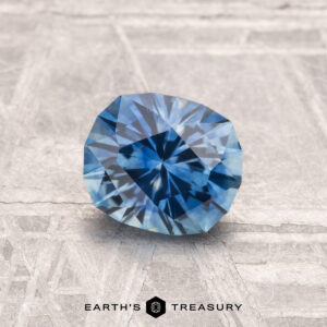 A 1.18-carat heated Montana sapphire, cut in a faceted oval shape, rests on a textured, light-colored surface with "Earth's Treasury" written below.