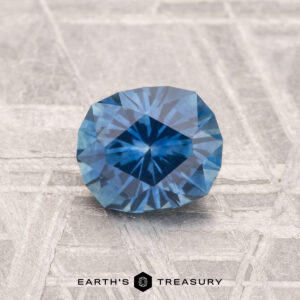 A 1.15-carat heated Montana sapphire with a cushion cut rests on a textured surface, with the text "EARTH'S TREASURY" below.