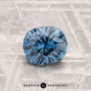 A 1.13-carat Montana sapphire (heated) is displayed on a textured gray surface, with the text "EARTH'S TREASURY" at the bottom.