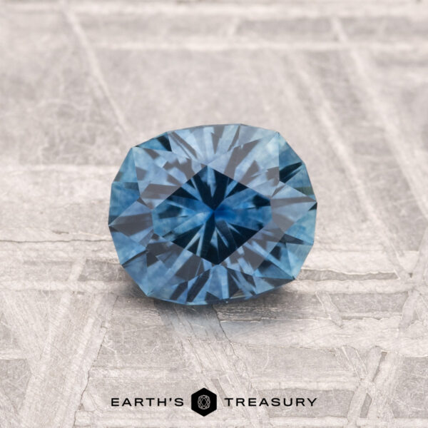 1.13-Carat Montana Sapphire (Heated) A 1.13-carat Montana sapphire (heated) is displayed on a textured gray surface, with the text "EARTH'S TREASURY" at the bottom.