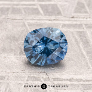 A 1.09-carat heated Montana sapphire is displayed on a textured gray surface with the words "EARTH'S TREASURY" printed below.