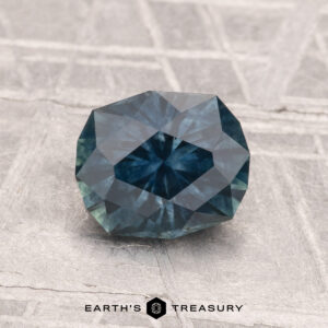 A 1.51-carat heated Montana sapphire with a faceted, dark bluish-green hue rests on a textured gray surface, featuring the "EARTH'S TREASURY" logo and text at the bottom.