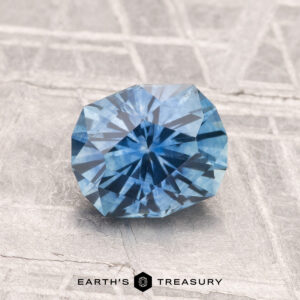 A 1.42-carat heated Montana sapphire is displayed on a textured gray surface with the text "Earth's Treasury" at the bottom.
