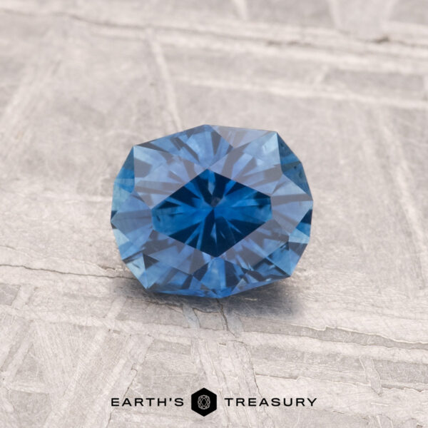 0.94-Carat Montana Sapphire (Heated) A 0.94-carat heated Montana sapphire with a faceted geometric cut rests on a textured gray surface, with "Earth’s Treasury" appearing at the image’s bottom.
