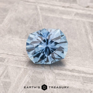A round, faceted 0.77-carat heated Montana sapphire with a blue hue is displayed on a light textured surface, featuring the text "Earth's Treasury" at the bottom.