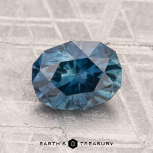 A 2.20-carat heated Montana sapphire, faceted in an oval shape, rests on a textured metallic surface displaying "EARTH'S TREASURY" at the bottom.