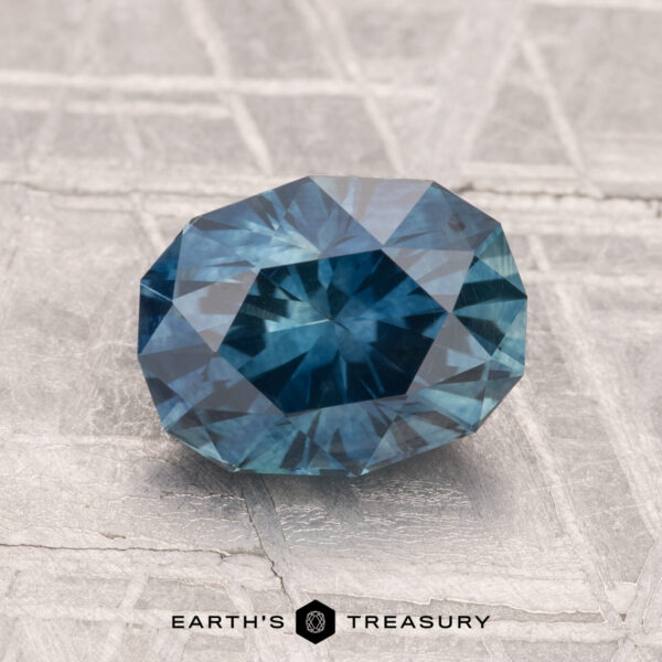 2.20-Carat Montana Sapphire (Heated) A 2.20-carat heated Montana sapphire, faceted in an oval shape, rests on a textured metallic surface displaying "EARTH'S TREASURY" at the bottom.