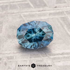 A 1.31-carat heated Montana sapphire, cut in a faceted oval shape, is displayed on a textured gray surface with "EARTH'S TREASURY" at the bottom.
