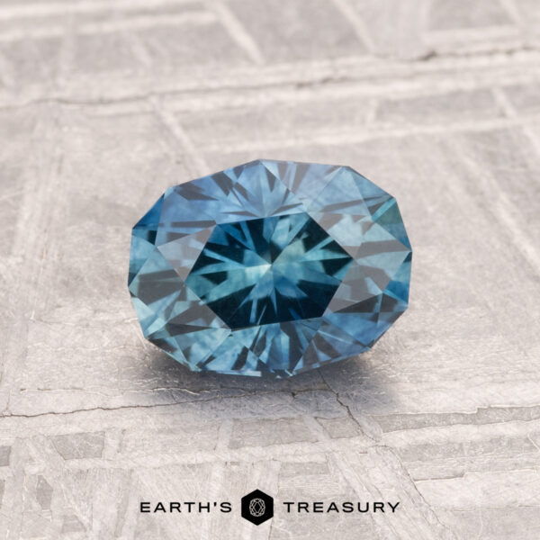 1.31-Carat Montana Sapphire (Heated) A 1.31-carat heated Montana sapphire, cut in a faceted oval shape, is displayed on a textured gray surface with "EARTH'S TREASURY" at the bottom.