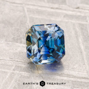 A 1.19-carat heated Ceylon sapphire with blue-green hues sits on a light, textured surface, featuring the "Earth's Treasury" text at the bottom.