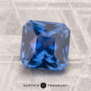 A 2.03-carat heated Ceylon sapphire, cut in a square shape, rests on a textured gray surface with "Earth's Treasury" displayed below.