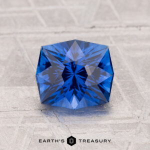A 1.75-carat heated Ceylon sapphire, cut in a blue hexagonal facet, rests on a light textured surface with "EARTH'S TREASURY" written below.